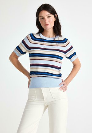 Donna con capelli scuri che indossa un maglione azzurro a maniche corte con righe orizzontali multicolori e pantaloni bianchi, in piedi con le mani sui fianchi.