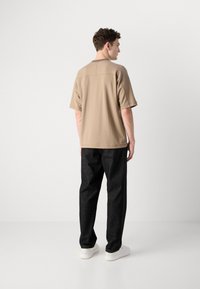 Beige short-sleeve shirt with a straight fit, detailed stitching, paired with black denim trousers and white sneakers, viewed from the back.