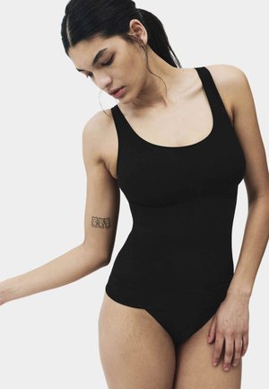 Frau trägt einen schwarzen ärmellosen Body, schaut nach unten, mit einem Tattoo von drei Teddybären auf ihrem oberen linken Arm.
