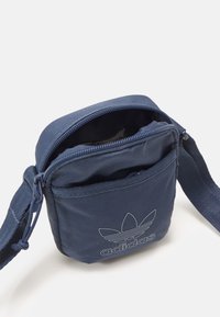 Marinblå tygpåse med dragkedjestängning, justerbar rem och framficka. Har vit Adidas-logotyp präntad på framsidan.