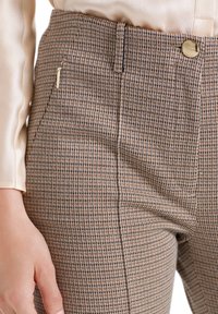 Pantalon marron à motif pied-de-poule avec une texture lisse, présentant une main posée sur la hanche et une fermeture éclair accentuée en or sur le côté.