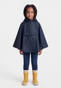 Mlada djevojka s pletenicama, obučena u mornarskoplavi kišni pončo, tamne traperice i žute kišne čizme, stoji ispred bijele pozadine.