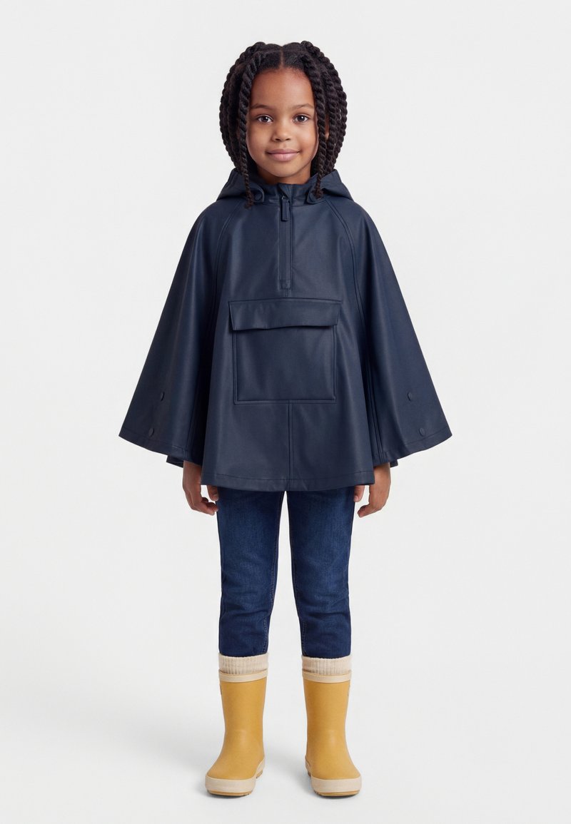 Mlada djevojka s pletenicama, obučena u mornarskoplavi kišni pončo, tamne traperice i žute kišne čizme, stoji ispred bijele pozadine.