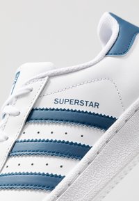 White sneaker with blue accents, featuring three horizontal stripes, textured overlays, and a rubber sole. "SUPERSTAR" printed on the side.