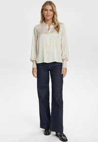 Blusa beige chiaro con bottoni frontali, dotata di maniche lunghe e arricciate. Abbinata a jeans a gamba larga di un blu scuro e scarpe nere.