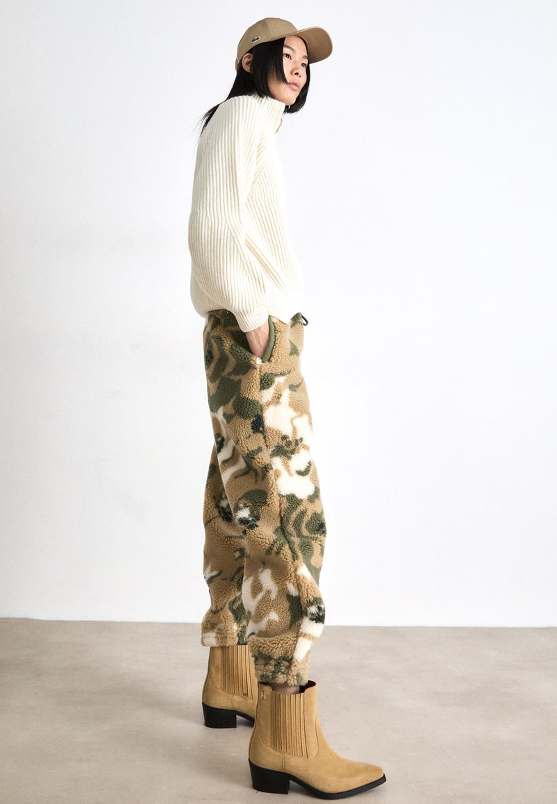 Kremowy sweter w prążki, wzorzyste brązowe i zielone spodnie polarowe oraz beżowe botki za kostkę z frędzlami. Na głowie czarna czapka.