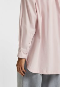 Light pink long-sleeve shirt with a buttoned cuff, made of smooth fabric; features subtle stitching detail and a relaxed silhouette.