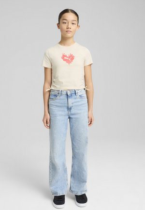 Camiseta cropped beige con un gráfico de corazón de llamas rojas, combinada con jeans de pierna ancha de color azul claro. El look presenta cordones en los lados de la camiseta.