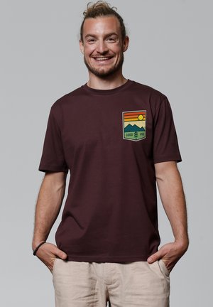 Bordeaux-kleurig T-shirt met korte mouwen, voorzien van een grafische afbeelding op de linkerborst met een zonsondergang, bergen en bomen, gedrukt in groen, oranje en geel.