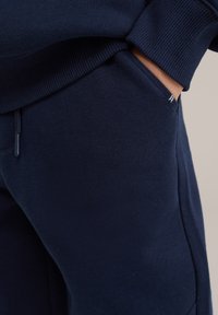 Tumši zila kokvilnas maisījuma sporta bikses ar ribotām jostām un aprocēm. Iespējo sānu kabatu un mazu logotipa uzlīmi uz jostas.