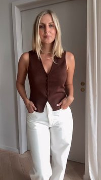 Ruda megzta besiūlė palaidinė su V formos iškirpte ir priekyje užsegamais mygtukais, derinama su aukšto juosmens baltais kelnais, prieš neutralią foną.