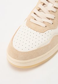Beige Wildledersneaker mit weißen Lederakzenten, gestanzten Löchern für Atmungsaktivität und cremefarbenen Schnürsenkeln. Strukturierte Seiten und Gummisohle.