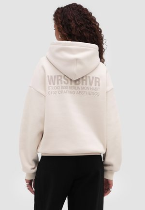 Vrouw met krullend haar draagt een crème kleurige hoodie met de capuchon op, zwarte broek, toont tekst "WRSTBHVR STUDIO (030) BERLIN MON HABIT 0102 CRAFTING AESTHETICS" op de achterkant.