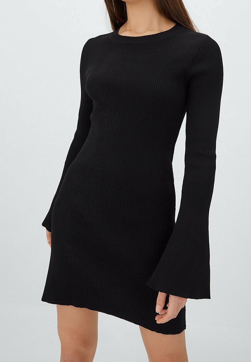 Femme portant une robe noire cintrée en maille côtelée avec des manches longues évasées et un col rond, debout devant un fond clair uni.