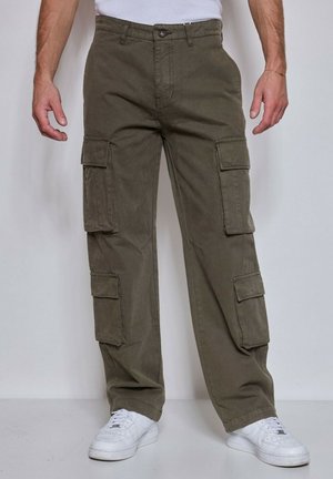 Homme portant un pantalon cargo vert olive avec plusieurs poches et des baskets blanches, debout devant un fond uni.