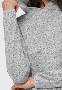 Sweat à capuche texturé gris avec un col haut, doté de manches longues et d'un tissu tricoté doux. Le matériau présente un motif tacheté.