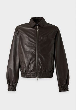 Jeune homme portant une veste en cuir marron foncé, une chemise beige à carreaux, un pantalon gris ample et des baskets noires, debout devant un fond blanc uni.