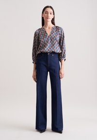 Knopfbluse mit geometrischem Muster in Blau, Rot und Creme, kombiniert mit hochtaillierten weit geschnittenen Jeanshosen in Dunkelblau.