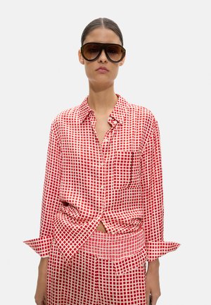 Kobieta nosząca duże, ciemne okulary przeciwsłoneczne oraz czerwoną i białą koszulę w kratę zapinaną na guziki, z dopasowanymi spodniami.