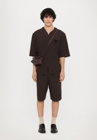Brown short-sleeve button-up shirt with embroidered "W" and matching shorts. Accessories include a small brown crossbody bag and beige socks.