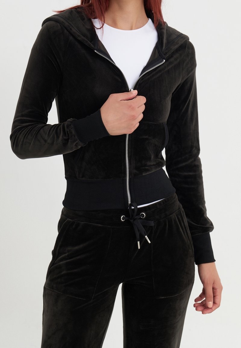 Femme portant un sweat à capuche zippé en velours noir et un pantalon assorti, avec un t-shirt blanc en dessous, main tenant la fermeture éclair au niveau du milieu de la poitrine.