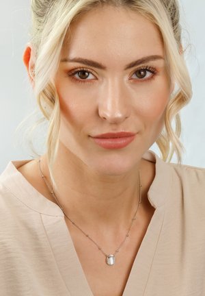 Silver necklace with a small, oval pendant, delicate chain featuring round accents, worn over a light beige blouse.
