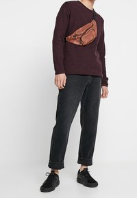 Suéter marrón, jeans de mezclilla negros, zapatillas negras y una riñonera de cuero marrón con dos cremalleras. Ajuste casual y diseño relajado.