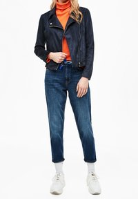 Giubbotto da moto in suede blu navy con design aderente, zip asimmetrica e un dolcevita arancione sotto. Abbinato a jeans blu a vita alta.