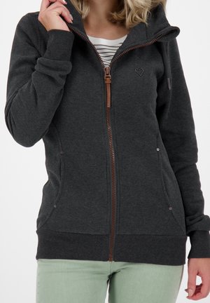 Femme portant un sweat à capuche gris foncé avec un petit détail en forme de cœur, partiellement dézippé sur une chemise blanche à rayures et un pantalon vert clair.