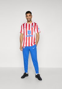 Futbalový dres červeno-biely pruhovaný s modrými akcentmi, skombinovaný s modrými teplákmi a čiernymi športovými topánkami. Viditeľné logo Nike.