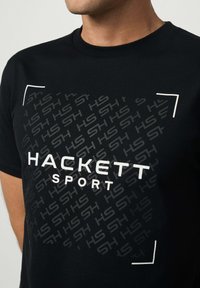Musta t-paita, jossa on viiston toistuvan "HS" -kuvion ja rohkeat valkoiset tekstit "HACKETT SPORT" valkoisten kulmalevyjen keskellä.