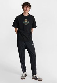 Svart bomullst-shirt med en grön och guldfärgad grafikdesign, kombinerad med svarta träningsbyxor med logotyp och kontrasterande grå sidpaneler.