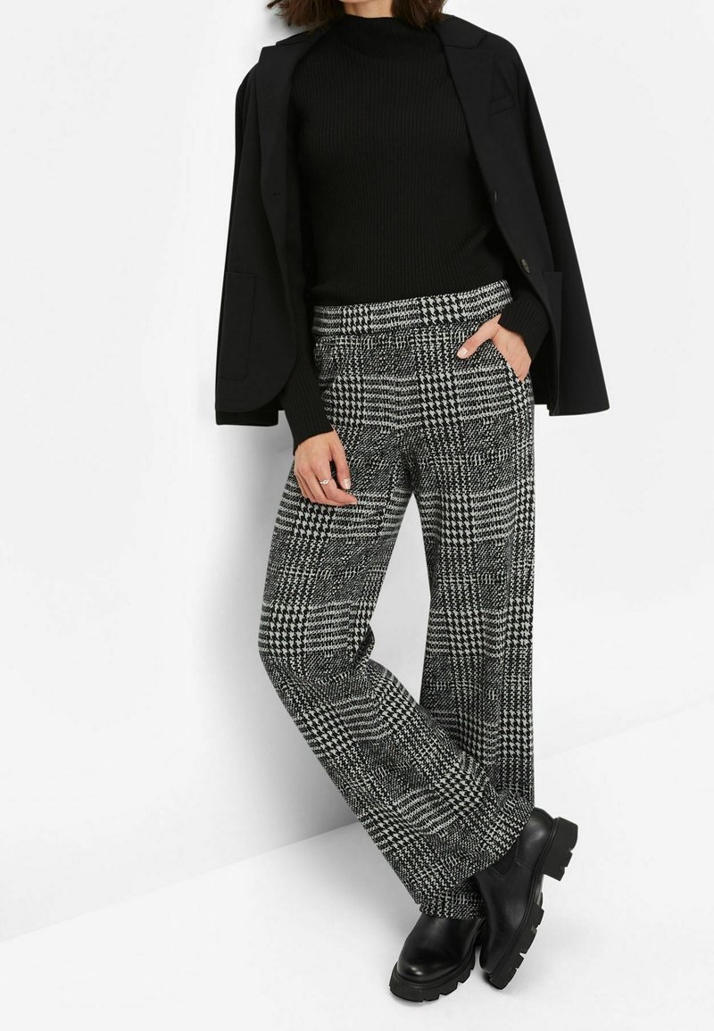 Femme portant un blazer noir, un pull côtelé noir, un pantalon à carreaux pied-de-poule noir et blanc, ainsi que des bottes noires épaisses, debout contre un mur blanc.