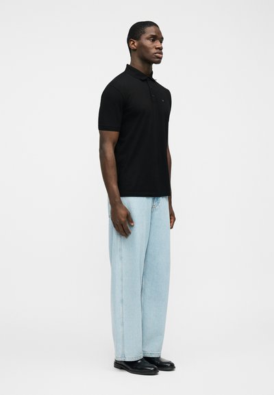 Άνδρας που στέκεται και βλέπει προς τα δεξιά, φοράει μαύρο πόλο με κοντά μανίκια, ανοιχτό μπλε τζιν παντελόνι με φαρδιά πόδια και μαύρα παπούτσια σε λευκό φόντο.