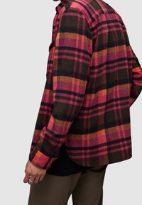 Koszula flanelowa w kratę w kolorze różowym, pomarańczowym i brązowym z czarnymi akcentami; ma długie rękawy, zapięcie na guziki i zaokrąglony dół.