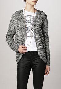 Černobílý vzorovaný cardigan s otevřeným předním designem, pletená textura, dlouhé rukávy, kombinovaný s grafickým tričkem a černými kalhotami.