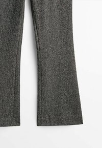 Gros plan sur des jambes de pantalon en tissu gris avec un motif chevrons noir et blanc subtil sur fond blanc.