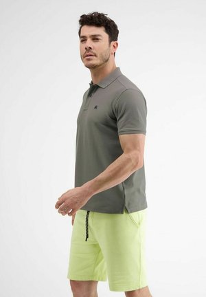 Mann trägt olivgrünes Poloshirt und hellgrüne Shorts, steht seitlich vor einem schlichten weißen Hintergrund.