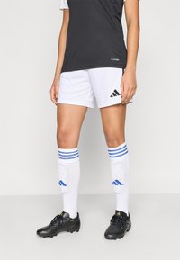 Μαύρη και λευκή αθλητική στολή που περιλαμβάνει κοντομάνικη μπλούζα, λευκό σορτς με μαύρες λεπτομέρειες, κάλτσες με μπλε ρίγες και μαύρα παπούτσια με τακούνια.