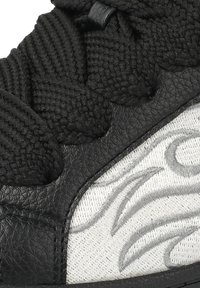 Gros plan sur des lacets tissés noirs sur du cuir noir et un tissu blanc avec des volutes brodées grises sur une basket.