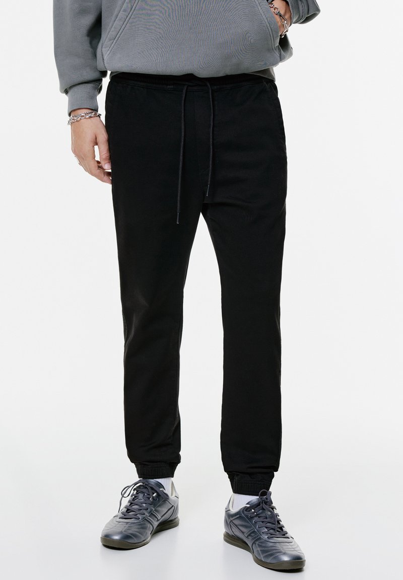 Zwarte joggers met een elastische tailleband en een trekkoorddetail. Ze hebben gebundelde enkels en een gladde, textuuroverfinish.