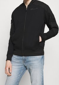 Man med en svart zip-up bomberjacka med ribbade manschetter och ljusblå urtvättade jeans, med en hand i jackfickan.