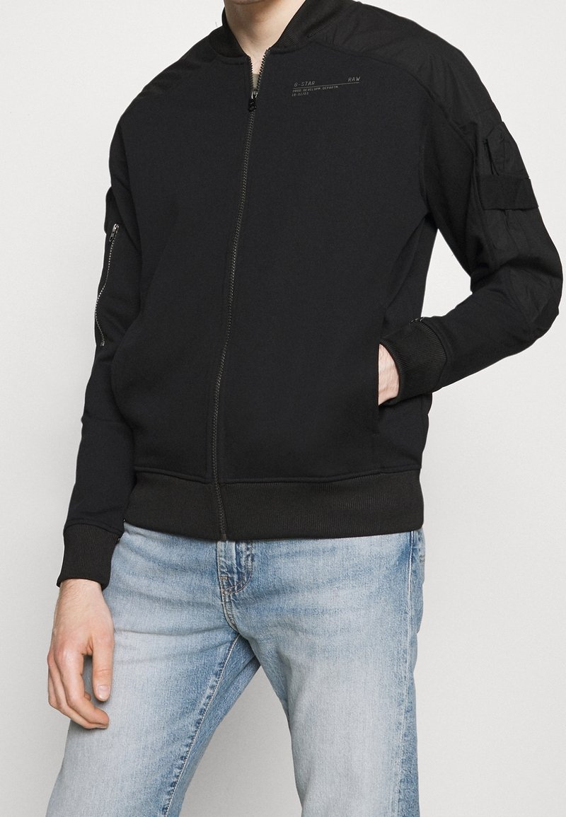 Man med en svart zip-up bomberjacka med ribbade manschetter och ljusblå urtvättade jeans, med en hand i jackfickan.