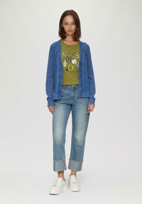 Cardigan bleu tricoté avec des motifs texturés, T-shirt vert avec impression graphique, jeans cropped bleu clair avec ourlets retroussés, baskets blanches.
