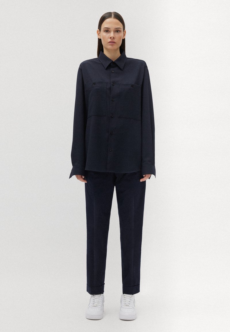 Marineblaues Hemd mit Knopfleiste und zwei Brusttaschen, kombiniert mit passenden schmal geschnittenen Hosen und weißen Sneakers, mit einer glatten Stoffoberfläche.