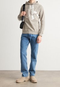 Sudadera gris con gráfico blanco, jeans azules y botas beige sin cordones. El modelo también sostiene un pequeño bolso cruzado negro. Texturas suaves visibles.