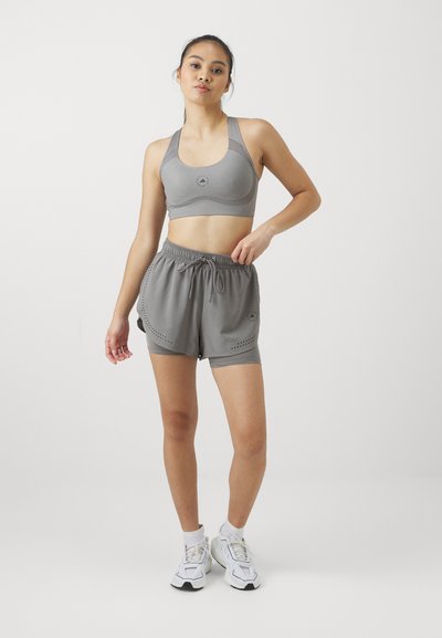 Sostén atlético gris con un logo redondo, combinado con pantalones cortos grises que tienen perforaciones laterales y una cintura con cordón. Zapatillas blancas completan el conjunto.