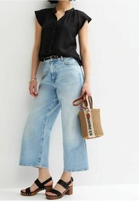 Blouse noire à manches courtes, jeans larges bleu clair, sandales à talons blocs noires, et un sac en paille tissé avec une inscription.