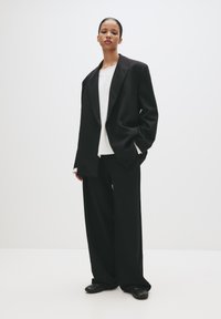 Femeie îmbrăcată într-un sacou negru supradimensionat, pantaloni largi negri, cămașă albă și pantofi negri, pe un fundal alb simplu.
