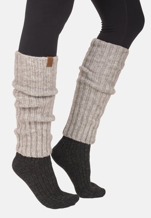 Benedenbenen met zwarte legging, donkergrijze geribbelde sokken en lichtgrijze grofgebreide beenwarmers op een witte achtergrond.